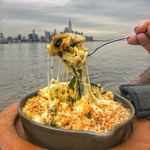 A hand holds a forkful of cheesy pasta with spinach, set against a city skyline by the water.