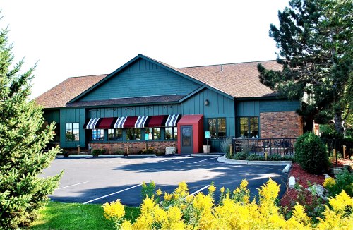 A green and brick building with large windows and a striped awning, surrounded by trees and colorful flowers.