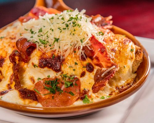 A close-up of a cheesy baked dish topped with crispy bacon, herbs, and grated cheese in a brown serving dish.