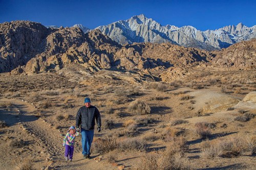 A person and a child walk hand in hand through a rocky desert landscape with snow-capped mountains in the background.