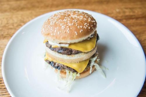 A stacked cheeseburger with two beef patties, melted cheese, lettuce, and a sesame seed bun on a white plate.