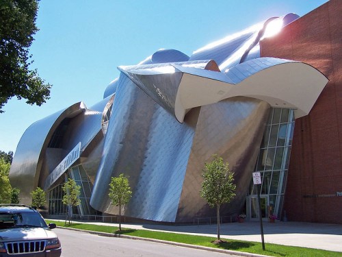 A modern, sculptural building with a metallic exterior and large glass windows, surrounded by trees and a clear blue sky.