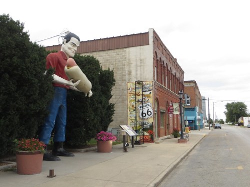 A giant statue of a man holding a sandwich stands beside a historic building on a street lined with plants and signs.