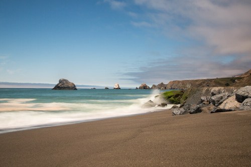 A serene beach scene with gentle waves, rocky formations, and a clear blue sky.