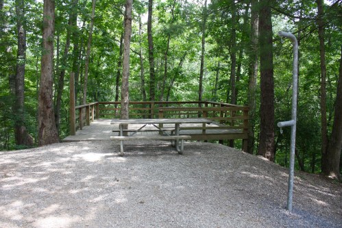 A wooden picnic platform surrounded by trees, with a gravel path and a metal post nearby.