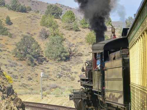A vintage steam train chugs along a scenic route, releasing black smoke against a backdrop of hills and trees.