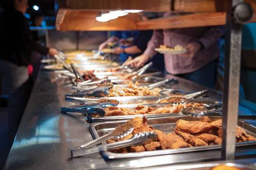 A buffet table with various dishes, people serving food with tongs under warm lighting.