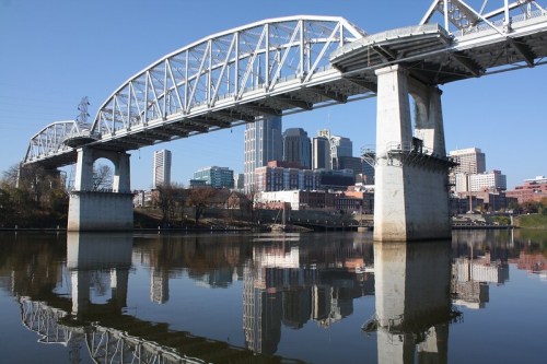 A bridge spans a river with city skyscrapers reflected in the water under a clear blue sky.