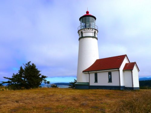 A white lighthouse with a red roof stands on a grassy area, surrounded by trees and a cloudy sky in the background.