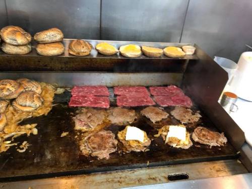 Griddle with cooking meats, melted cheese, and bagels on top, alongside bowls of condiments.