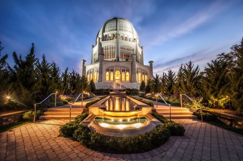 A beautifully lit temple at dusk, surrounded by lush greenery and a tranquil water feature in the foreground.