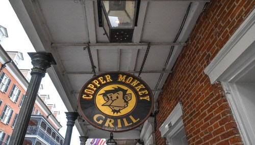 Sign for "Copper Monkey Grill" featuring a cartoon monkey, hanging from a porch awning with brick wall background.