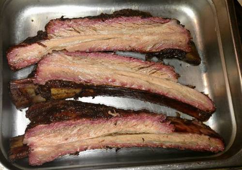 Four pieces of smoked beef ribs on a metal tray, showcasing a rich, dark crust and tender pink meat.
