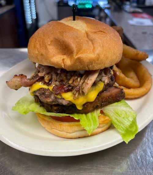 A hearty burger stacked with pulled pork, bacon, cheddar cheese, lettuce, and tomato, served with onion rings on the side.