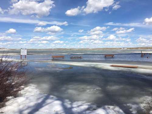 A flooded area with benches partially submerged, surrounded by ice and water under a blue sky with fluffy clouds.
