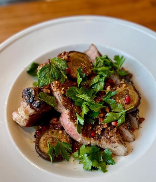 A plate of sliced meat topped with herbs, pomegranate seeds, and garnished with green leaves.