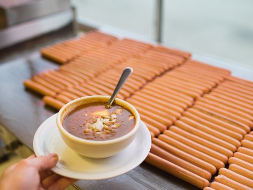 A hand holds a bowl of soup with a spoon, in front of a display of hot dogs.