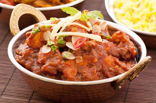 A bowl of rich, spicy chicken curry garnished with mint, sliced almonds, and red chilies, served with rice in the background.