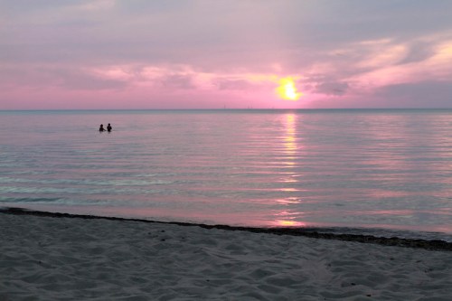 Two people wade in calm waters at sunset, with a pink and orange sky reflecting on the serene surface.
