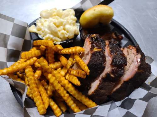 A plate of BBQ ribs, crinkle-cut fries, macaroni and cheese, and a biscuit, served on a checkered paper.