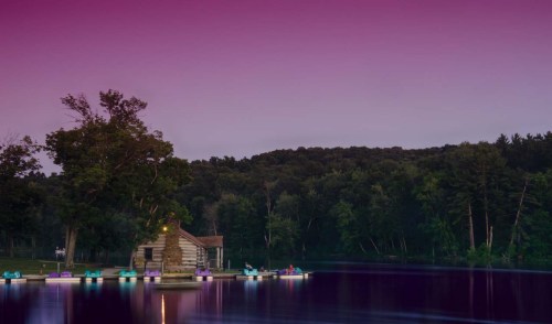 A serene lake at dusk with a rustic cabin and colorful lights reflecting on the water, surrounded by trees.