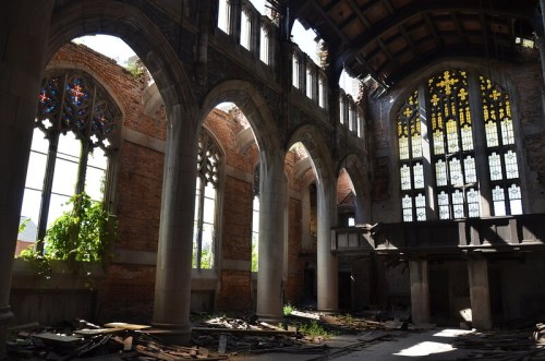 Interior of a dilapidated church with arched windows, exposed brick, and sunlight streaming through stained glass.