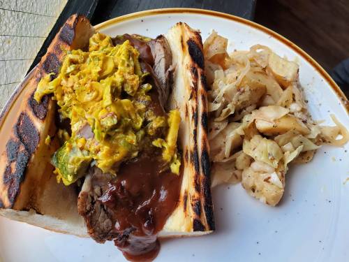 A toasted sandwich filled with brisket, topped with yellow sauce, served alongside sautéed cabbage and potatoes.