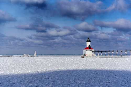 A lighthouse stands on a frozen shore under a cloudy sky, with ice covering the ground and water.