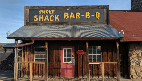 A rustic wooden building with a corrugated metal roof, featuring a sign that reads "Smoke Shack Bar-B-Q" and an open door.