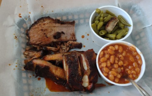 A plate with sliced smoked brisket, pulled pork, green beans, and baked beans in white cups.