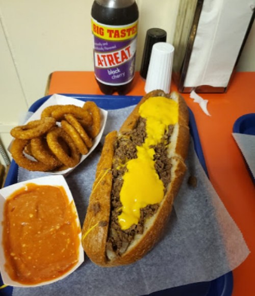 A tray with a cheesesteak sandwich, onion rings, and a cup of marinara sauce, alongside a bottle of cherry soda.