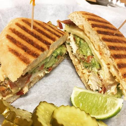 A sliced panini sandwich filled with chicken, avocado, and cheese, served with pickles and a lime wedge on the side.