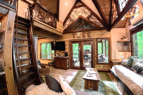 Cozy wooden cabin interior with a loft, large windows, and rustic decor, surrounded by greenery.