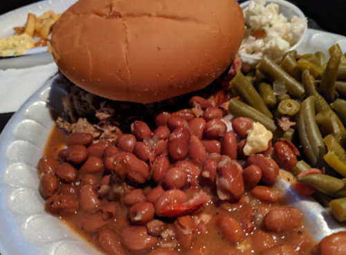 A plate featuring a pulled pork sandwich, pinto beans, green beans, and coleslaw on a white paper plate.
