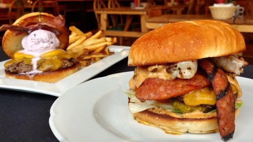 A close-up of a hearty burger with bacon, cheese, and toppings, alongside a plate of fries and a dessert burger.