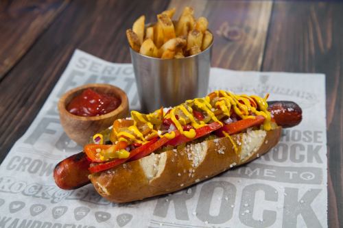 A gourmet hot dog topped with mustard, ketchup, and peppers, served with a side of fries in a metal cup.