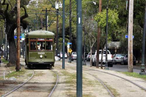 A green streetcar travels along tracks lined with trees and parked cars on a city street.