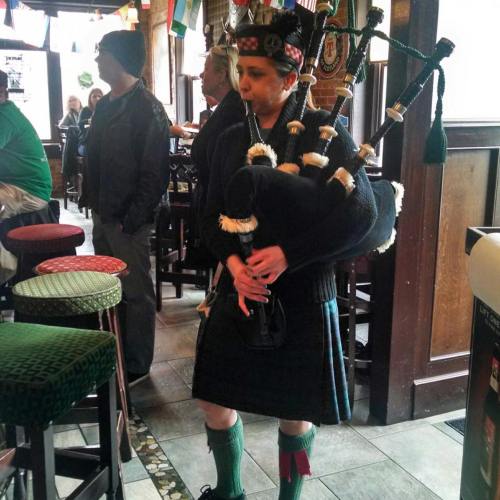 A person in a kilt plays the bagpipes inside a pub, surrounded by festive decorations and patrons.