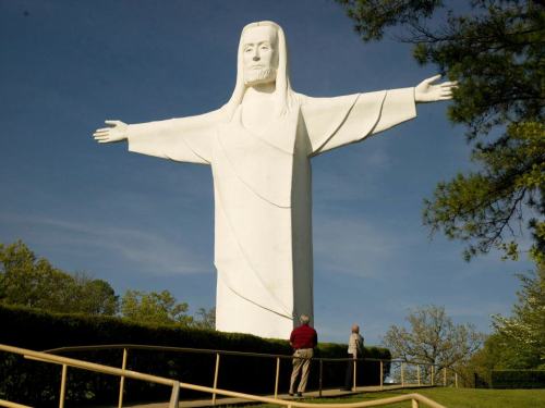 A large white statue of Christ with outstretched arms, surrounded by trees and two people standing nearby.