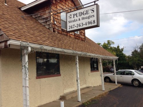 Exterior of Pudge's Steaks & Hoagies, featuring a sign and a building with a shingled roof and holiday lights.