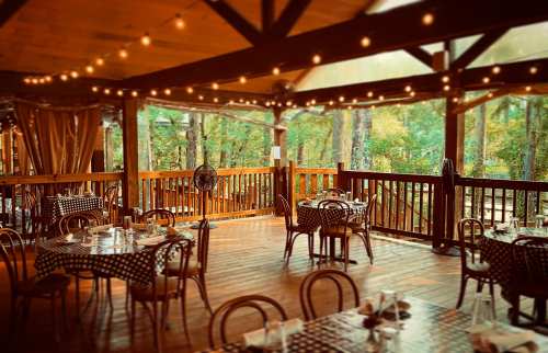 Cozy restaurant patio with checkered tables, wooden beams, and string lights, surrounded by trees.