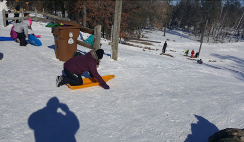 A person kneels on a bright orange sled on a snowy hill, with others sledding in the background.
