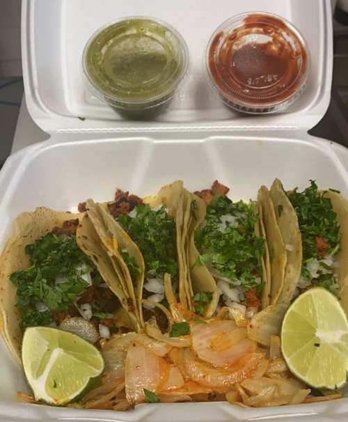 A takeout container with four tacos topped with cilantro and onions, served with lime wedges and two sauces.