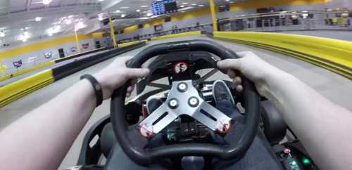 A driver's hands gripping a go-kart steering wheel, with a racetrack visible in the background.