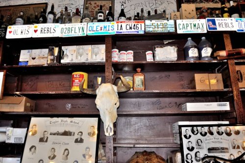 A wooden shelf filled with bottles, glasses, and memorabilia, featuring a cow skull and various license plates above.