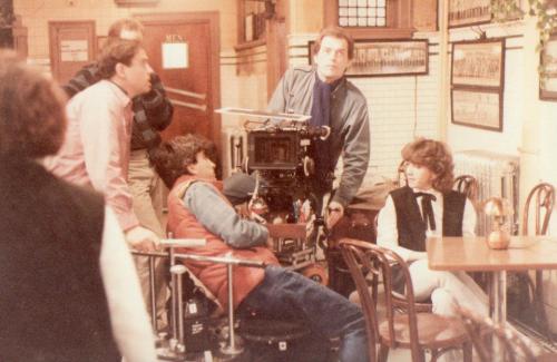 A film crew sets up a camera in a cafe, with actors and crew members engaged in discussion.