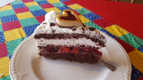 A slice of chocolate cake layered with whipped cream and cherries, topped with a heart-shaped decoration on a colorful tablecloth.