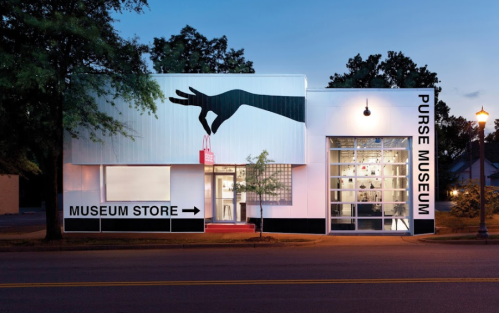 Exterior of the Purse Museum at dusk, featuring a large hand silhouette and a sign pointing to the museum store.
