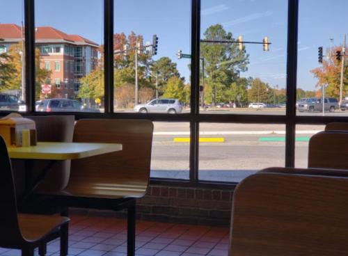 View from inside a restaurant, showing a sunny street with cars and trees outside through large windows.