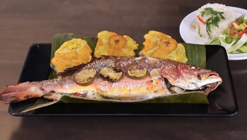 A whole grilled fish garnished with lime, served with fried plantains and a side of rice and salad.
